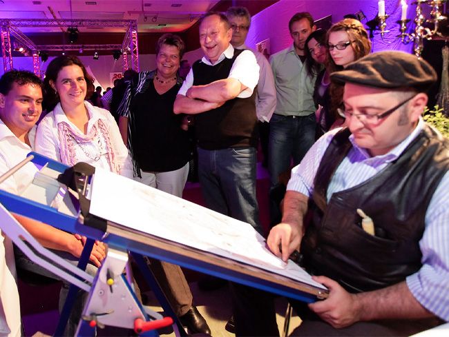 Ein junges Paar sitzt vor Erol, während er eine Karikatur zeichnet; umstehende Gäste lachen begeistert über die entstehende Zeichnung.