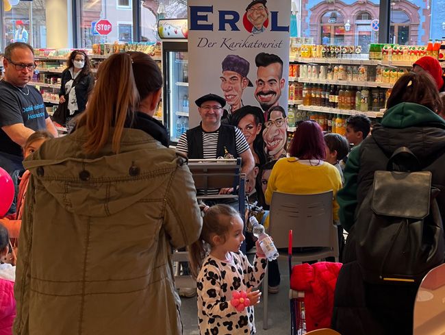 Erol zeichnet im Drogeriemarkt und ist von Kindern und Erwachsenen umringt, die für eine Karikatur Schlange stehen.