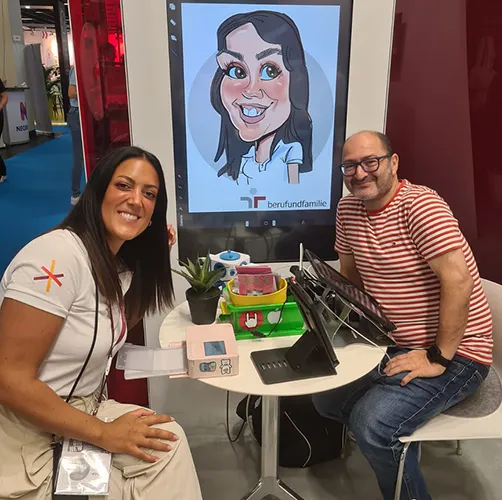 Erol zeichnet auf einer Messe in Köln eine digitale iPad-Karikatur für eine junge Frau, deren fertiges Porträt auf dem Bildschirm zu sehen ist.