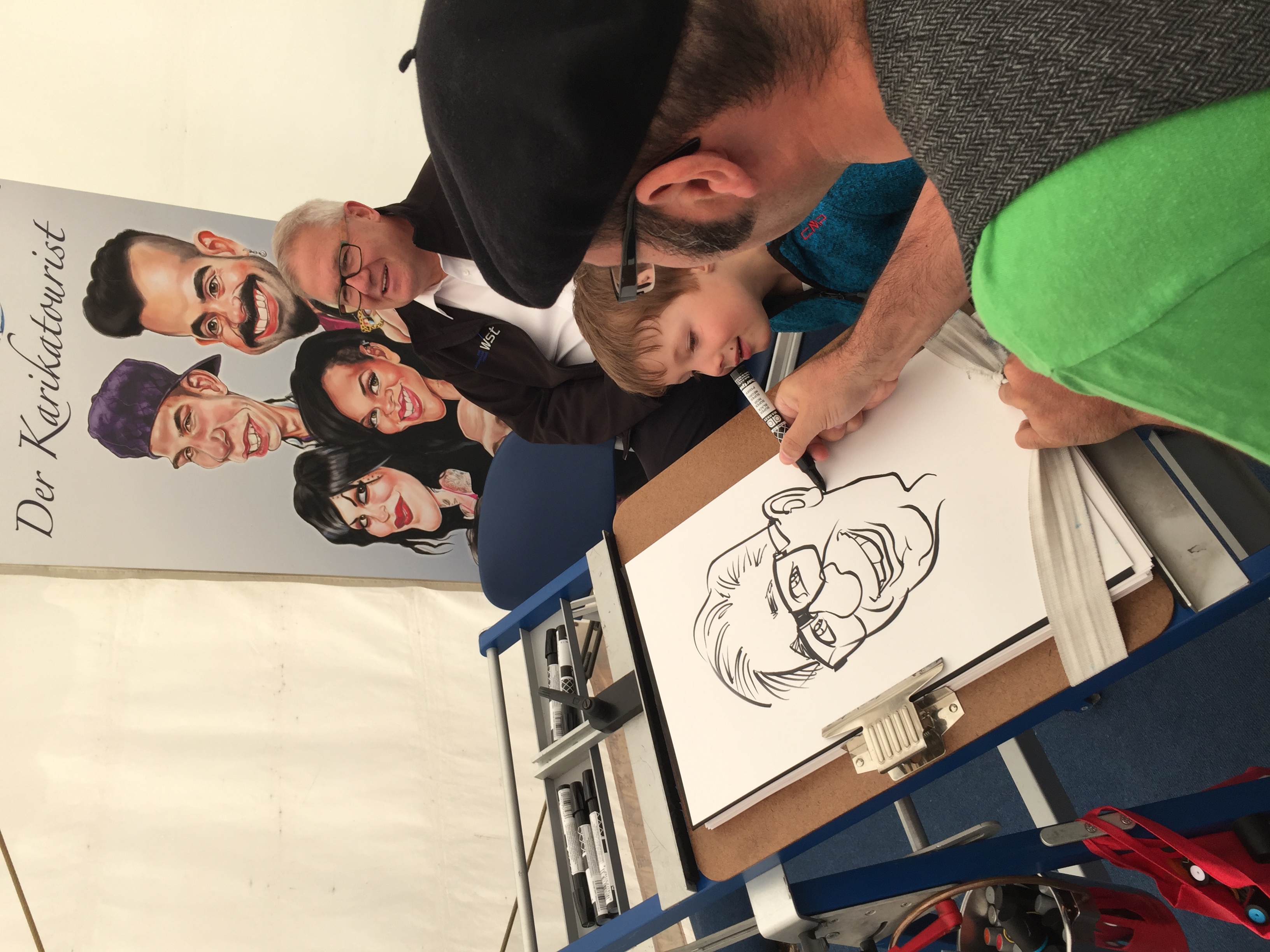 Erol zeichnet einen Herren, während ein junger Mann ihm aufmerksam zuschaut. Kunst und Kinderbetreuung in einem ;)